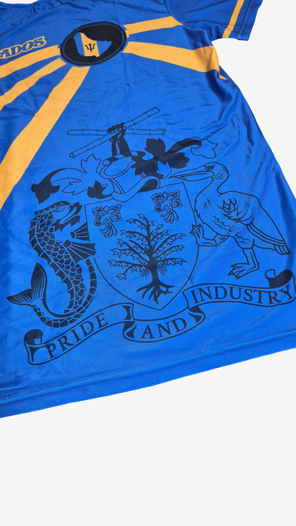 A close-up of a blue Barbados athletic jersey with yellow shoulder stripes and a large black crest graphic featuring a fish, bird, tree, and banner reading 'PRIDE AND INDUSTRY'.