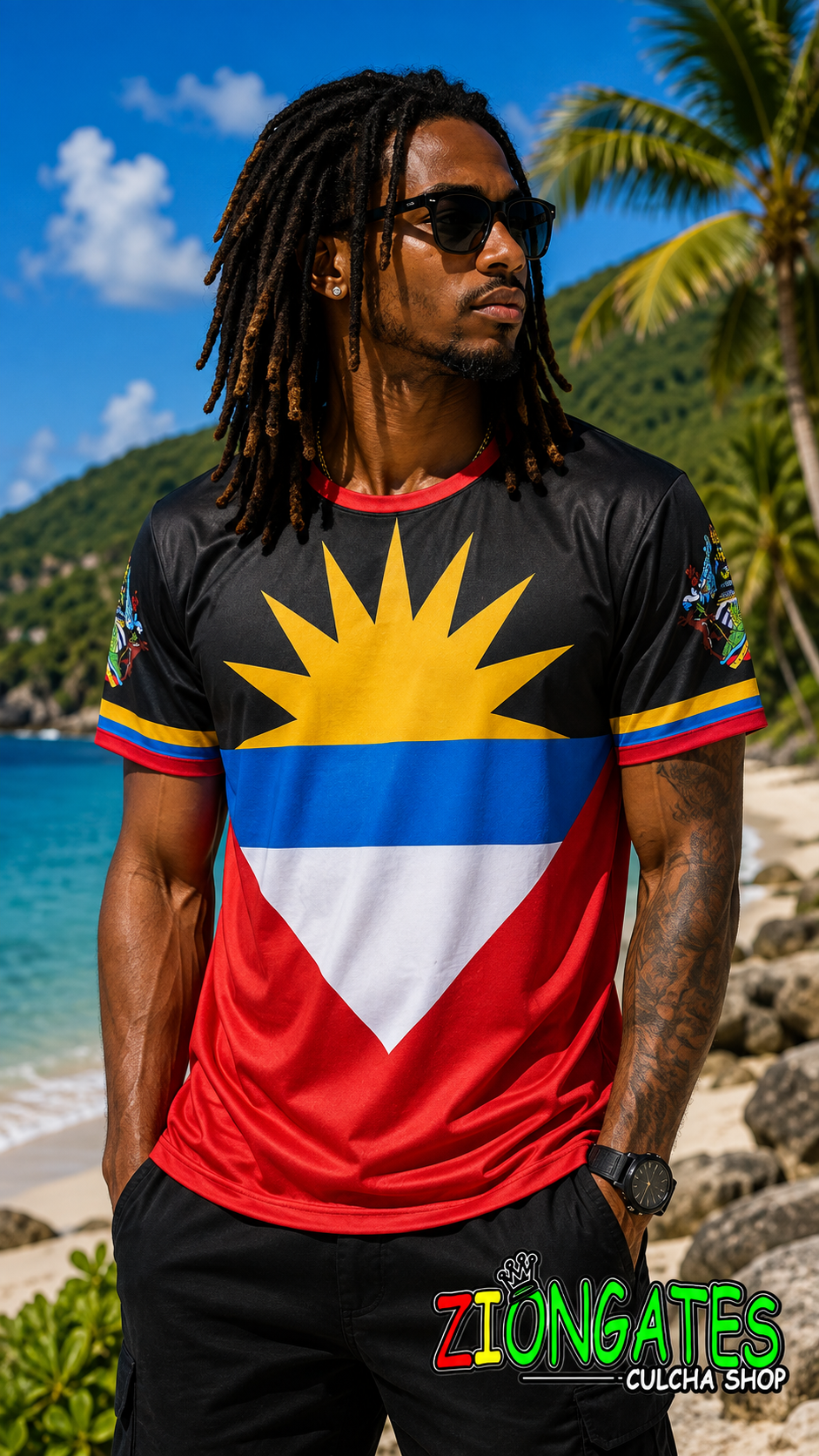 A man wearing a black athletic t-shirt with the flag of Antigua and Barbuda printed on the front, standing on a tropical beach with blue ocean and green hills in the background.